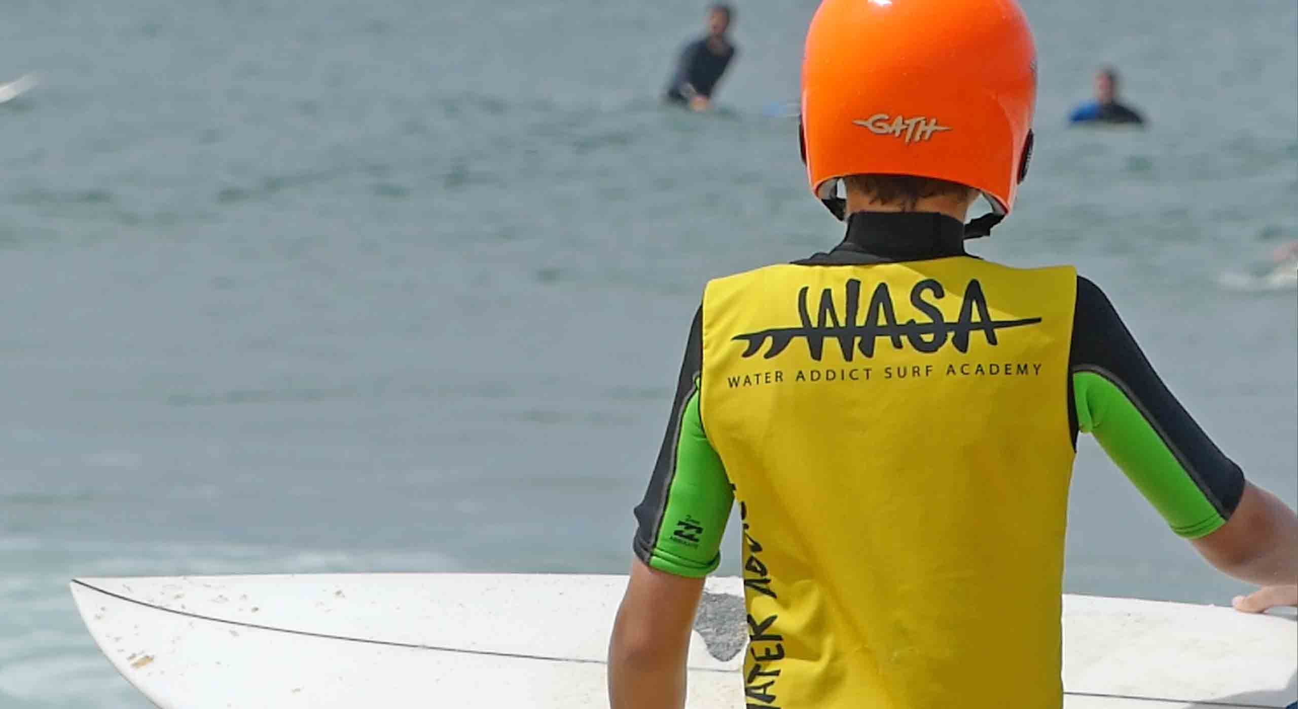 enfant de dos portant sa planche en résine avec un casque de surf orange et lycras jaune de l'école de surf wasa à capbreton